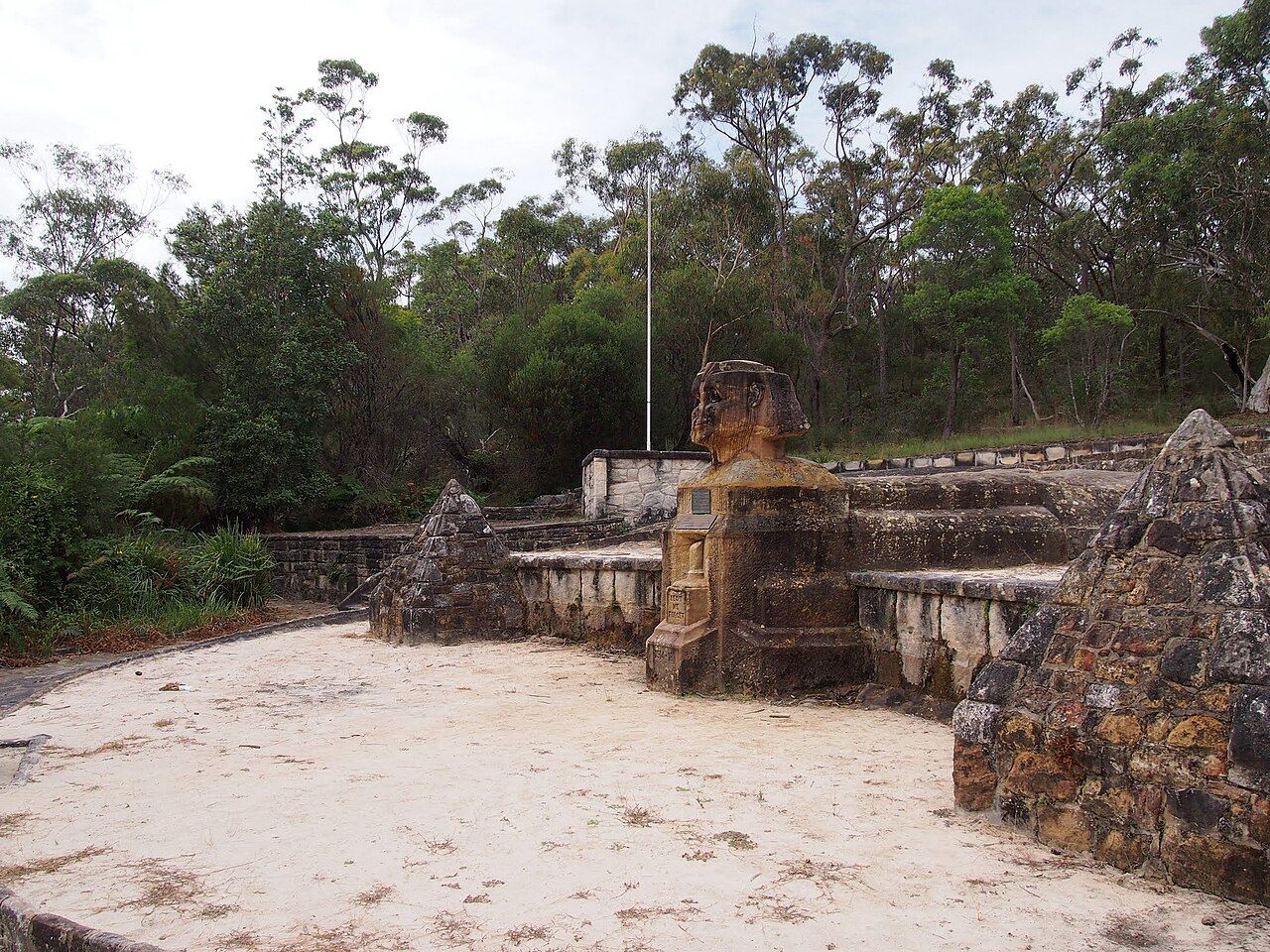 A carved sandstone sphinx on a plinth between two small stone pyramids, with bush behind